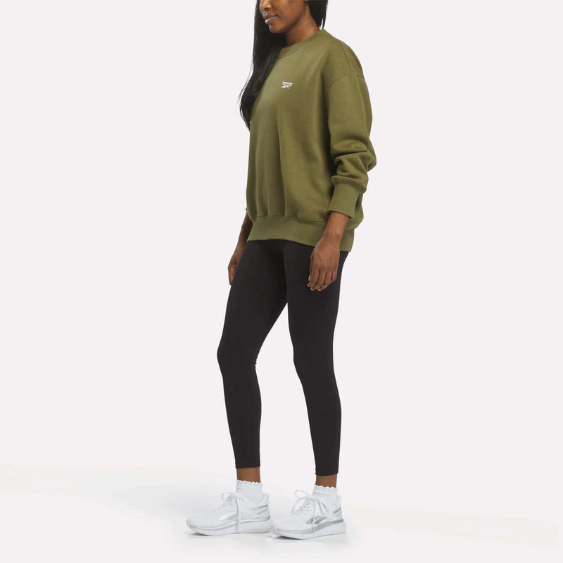 Woman in profile wears a Reebok Women’s Oversized Crewneck Sweatshirt in olive green, black leggings, and white sneakers, standing against a plain white background.