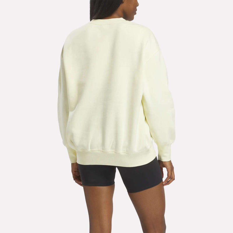 A person stands facing away from the camera against a plain background, wearing a Reebok Women’s Oversized Crewneck Sweatshirt in light yellow paired with black shorts.