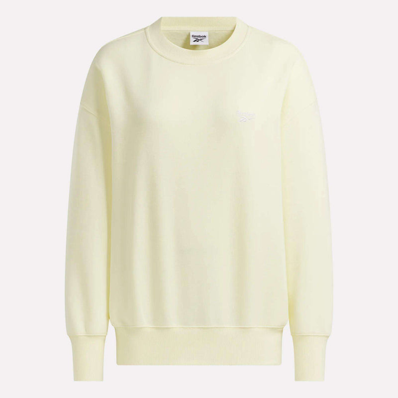 The Reebok Women’s Oversized Crewneck Sweatshirt is featured in light yellow, showcasing long sleeves, ribbed cuffs and hem, and a subtle white Reebok logo on the upper left chest. The sweatshirt is presented against a plain, light background.