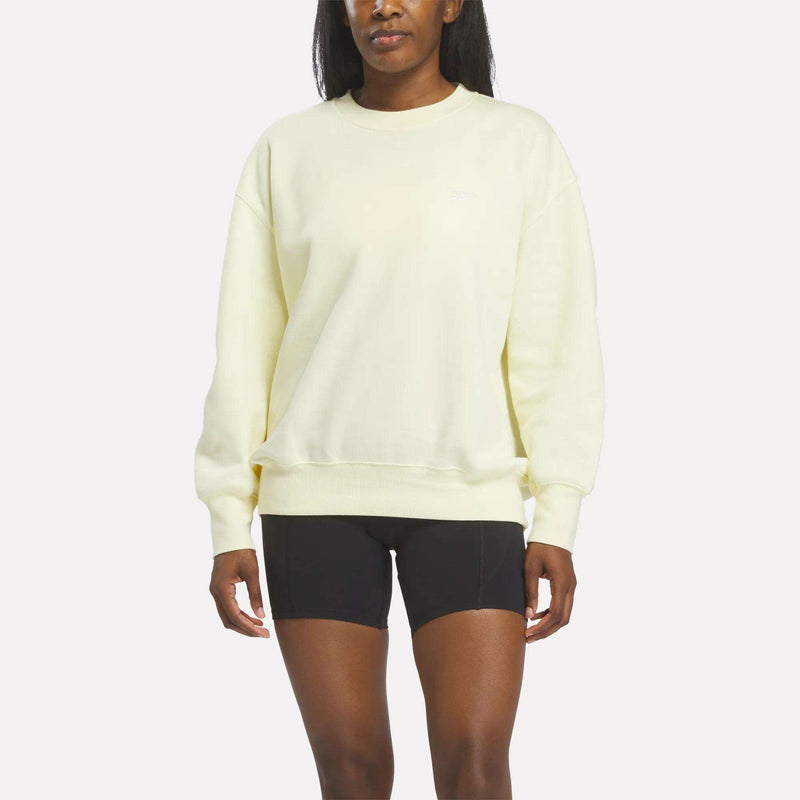 A person wearing a light yellow Reebok Women’s Oversized Crewneck Sweatshirt and black shorts stands against a plain white background, highlighting the comfortable design of the outfit.