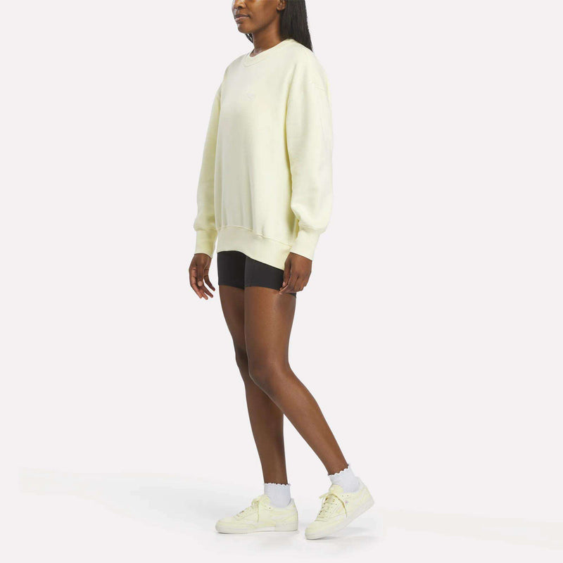 A person stands against a white background, wearing a Reebok Women’s Oversized Crewneck Sweatshirt in light yellow, black shorts, white socks, and light yellow Reebok sneakers.