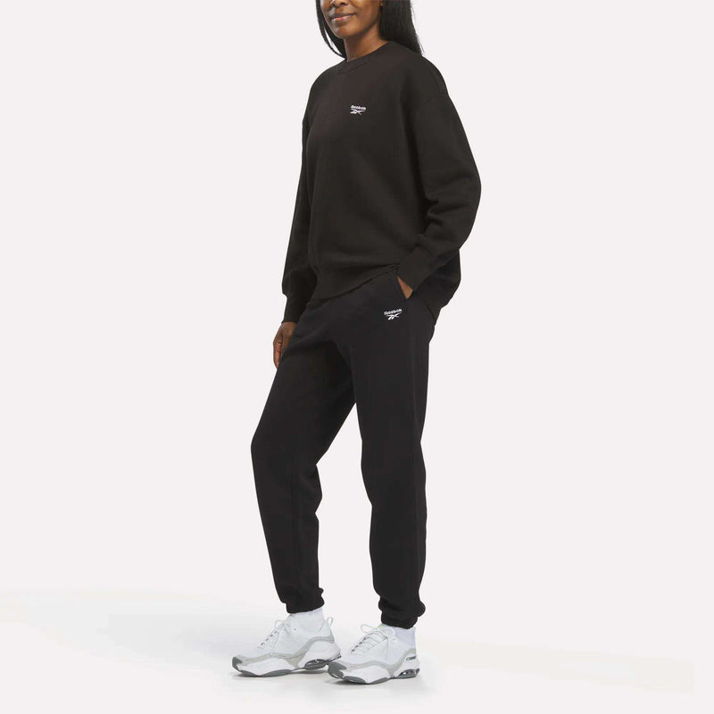 A person stands, showcasing the Reebok Women’s Cinch Sweatpants in black paired with a matching Reebok sweatshirt, both featuring subtle white logos. White sneakers complete the look, while the plain white background highlights the modern and comfortable style of this outfit.
