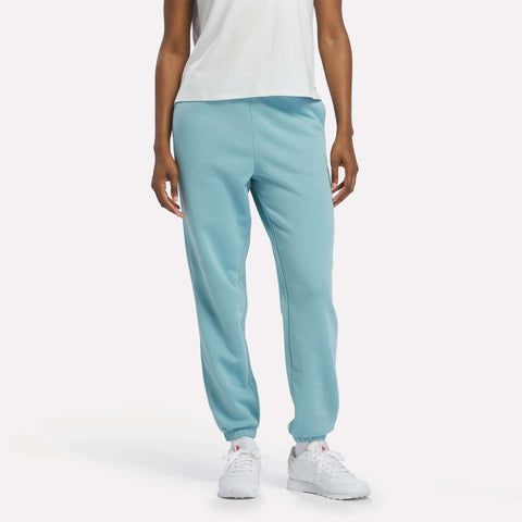 A person wears Women’s Cinched Sweatpants by Reebok in Hazy Teal, paired with a white short-sleeve shirt and white athletic shoes, posing against a plain light background. Only the body from shoulders down is shown.