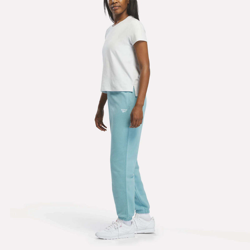 A woman stands sideways wearing a white t-shirt, Reebok Women’s Cinched Sweatpants in light blue, and white sneakers against a plain white background.