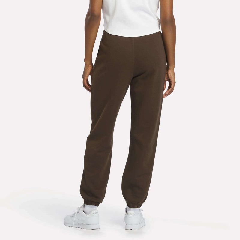 A person is standing facing away, wearing a white shirt, Reebok Women’s Cinch Sweatpants in dark brown with a modern design, and white sneakers against a plain background.