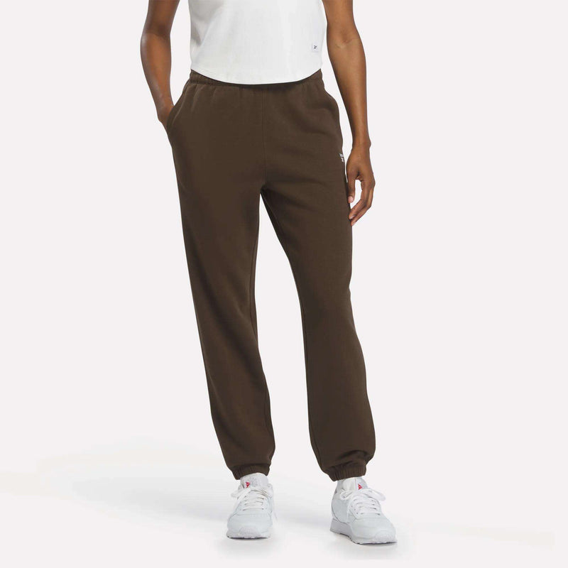 A person wearing a white shirt, Reebok Women’s Cinch Sweatpants in Espresso, and white sneakers stands against a plain white background with their hands in their pockets.