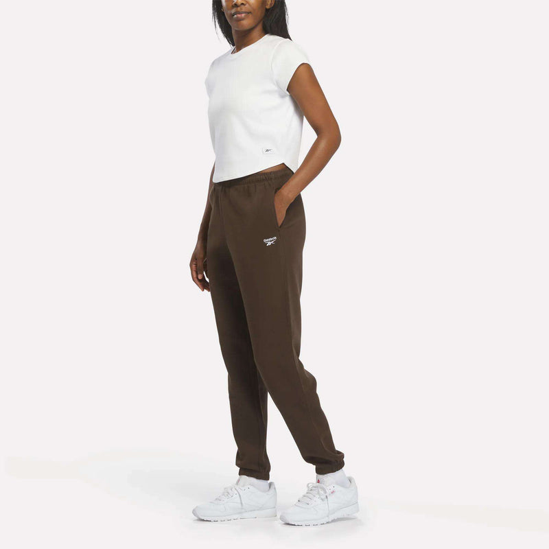 A woman stands with one hand in her pocket, wearing a white t-shirt, Reebok Women’s Cinch Sweatpants in brown, and white sneakers against a plain white background.