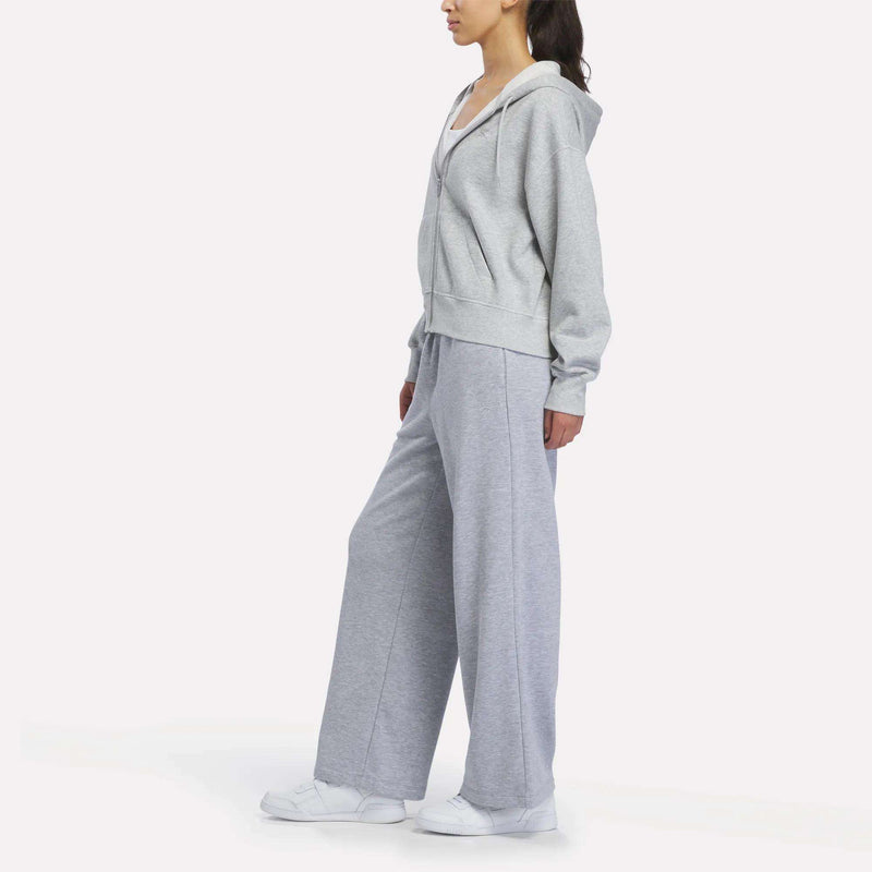A woman stands in profile against a plain white background, wearing Reebok Women’s Wide-Leg Sweatpants, a light gray hoodie, and white sneakers.
