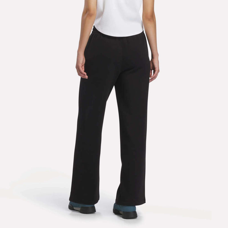A person stands facing away, wearing Reebok Women’s Wide-Leg Sweatpants in a relaxed fit, paired with a white sleeveless top and black shoes with blue accents, set against a plain light background.