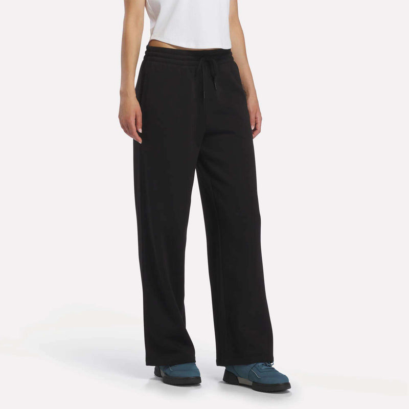 A person is shown from the lower torso down, wearing Reebok Women’s Wide-Leg Sweatpants in black, paired with a white T-shirt and blue shoes against a plain white background.
