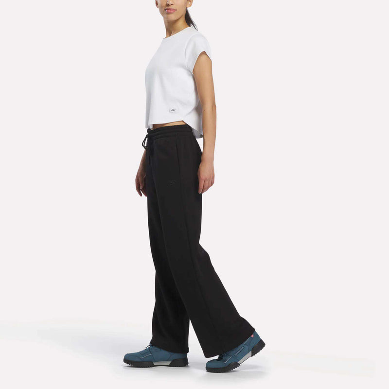 A person stands sideways against a plain white background, wearing a white short-sleeve shirt, Reebok Women’s Wide-Leg Sweatpants, and blue sneakers.