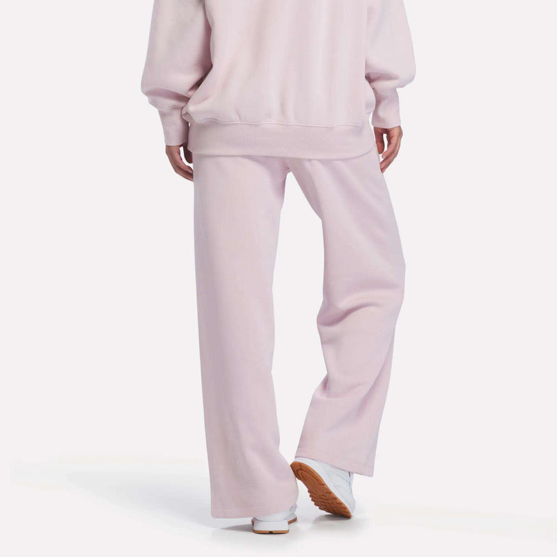 A person is shown from the back wearing Reebok Women’s Wide-Leg Sweatpants in light pink with a matching sweatshirt, paired with white sneakers that have brown soles.