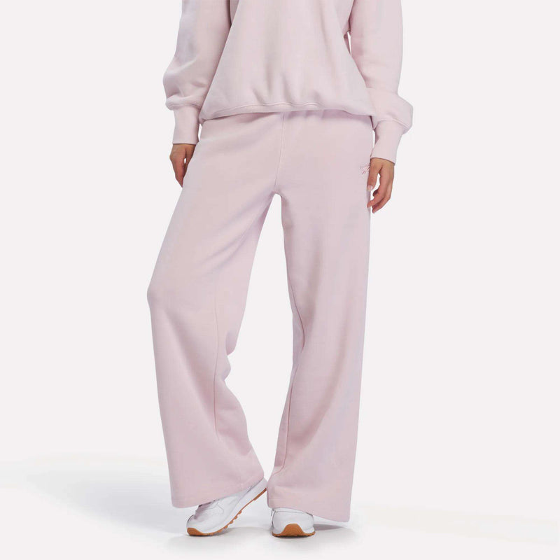 A person stands with one leg crossed over the other, modeling Reebok Women’s Wide-Leg Sweatpants in Elemental Pink along with the matching sweatshirt and white sneakers. The image is cropped from neck to feet on a white background.