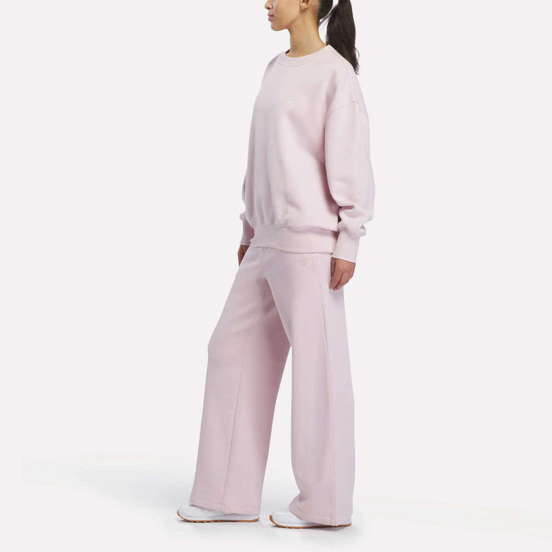 A person stands in a relaxed pose wearing pastel pink Women’s Wide-Leg Sweatpants from Reebok paired with an oversized sweatshirt and white sneakers, set against a plain white background. This effortlessly stylish look highlights the comfort and versatility of Reebok’s Women’s Wide-Leg Sweatpants while maintaining a chic, casual vibe.