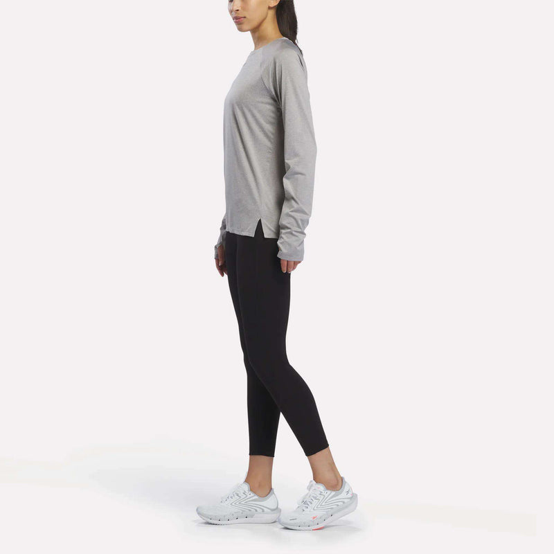 A woman stands sideways wearing a Reebok Women’s Long Sleeve Running Top, black leggings, and white sneakers against a plain white background.