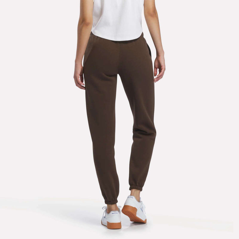 A person wearing a white sleeveless top, Reebok Women’s Logo Sweatpants in brown, and white sneakers is standing and facing away from the camera on a plain background.