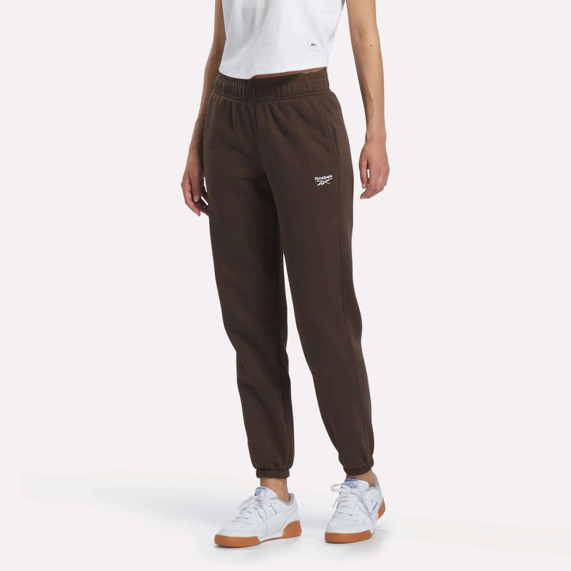 A person wearing a white T-shirt, Reebok Women’s Logo Sweatpants in espresso, and white sneakers stands against a plain background. Only the lower half of the body is visible, highlighting the sporty style of the sweatpants.