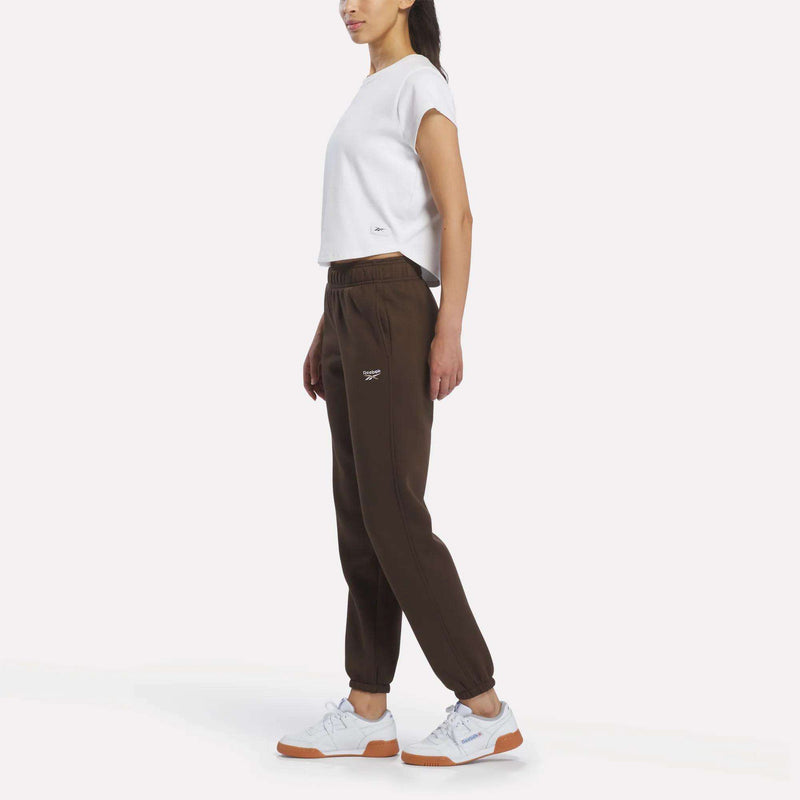 A woman stands sideways against a plain white background, wearing a white short-sleeve t-shirt and Reebok Women’s Logo Sweatpants in brown, paired with white sneakers featuring gum soles.