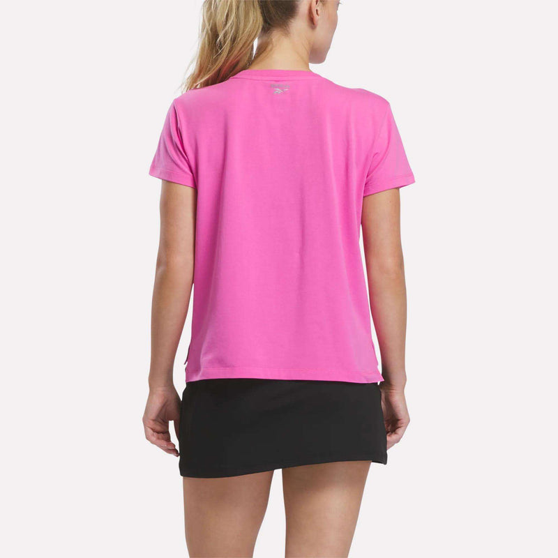A person with long blonde hair wears a bright pink short-sleeve shirt and a black Reebok Women’s Tennis Skort, standing with their back to the camera against a plain light background.