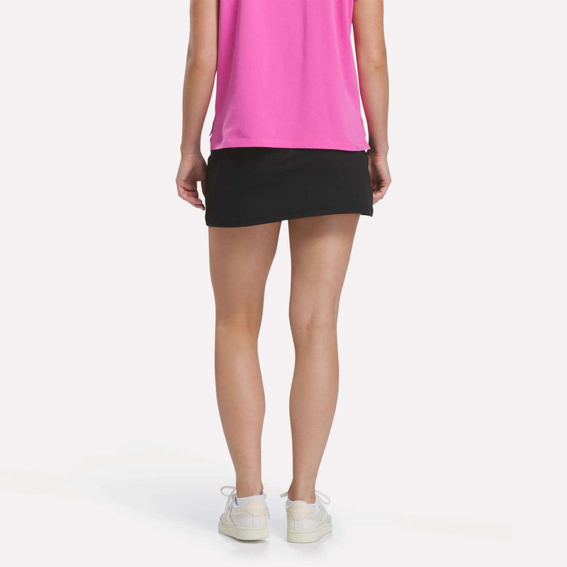 Person facing away, showing only the lower half, wears a bright pink shirt, white sneakers, and the Reebok Women’s Tennis Skort against a plain light background for court-inspired style.