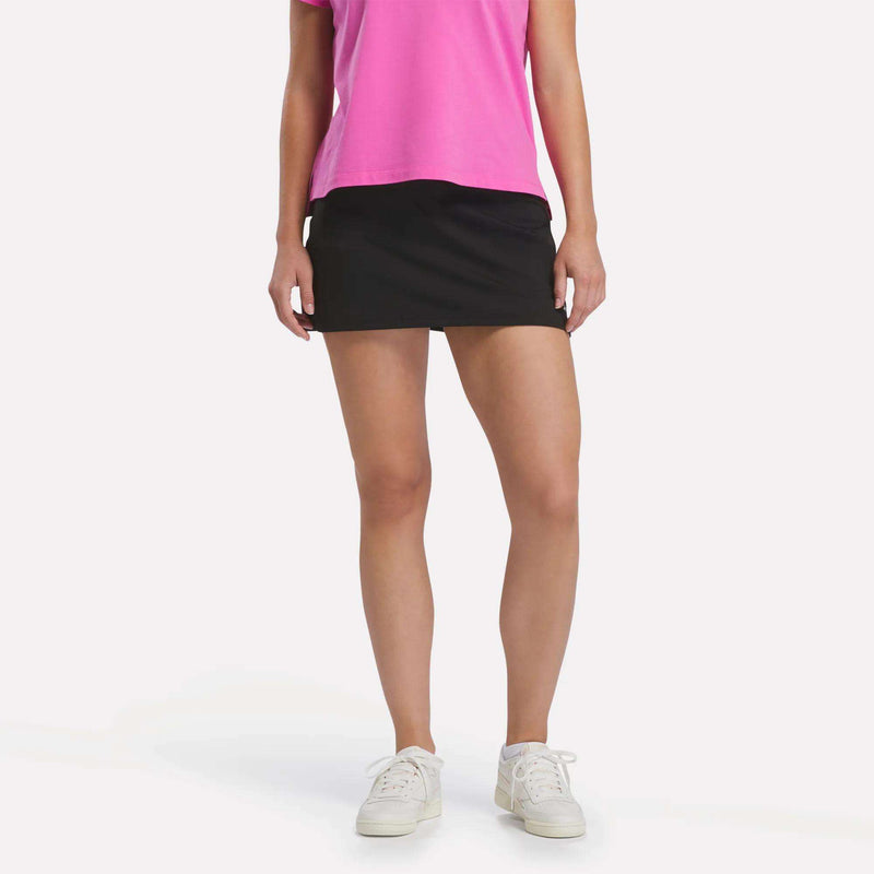 A person wears a pink t-shirt, Reebok Women’s Tennis Skort in black, and white sneakers, facing forward against a plain white background to showcase a court-inspired style.