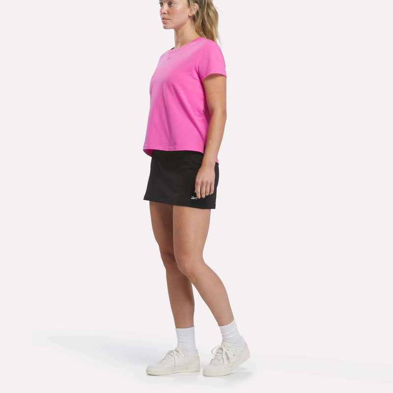 A woman wearing a black Reebok Women’s Tennis Skort, a pink T-shirt, white socks, and white sneakers stands against a plain white background, looking forward with a neutral expression.