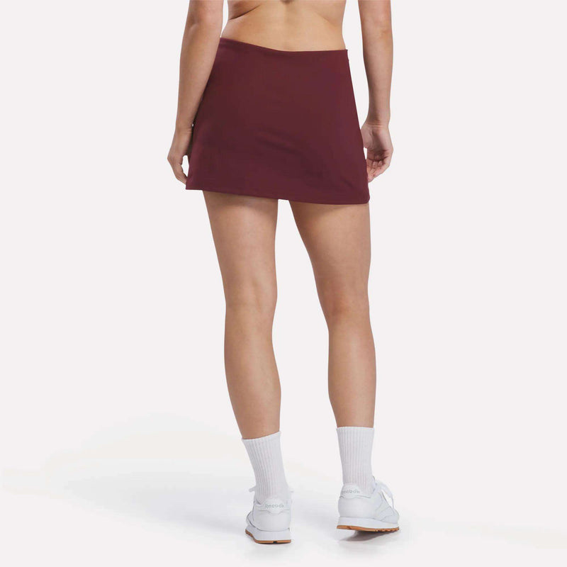 A person stands facing away, wearing the Reebok Women’s Tennis Skort in maroon, paired with white socks and white sneakers, set against a plain light background.