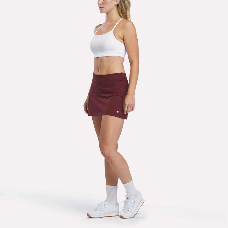A woman stands against a plain white background wearing a Reebok Women’s Tennis Skort with built-in shorts, paired with a white sports bra, white socks, and white sneakers.