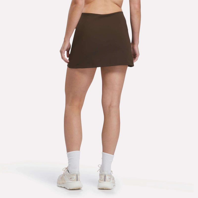 A person stands with their back to the camera, highlighting the Reebok Women’s Tennis Skort in dark brown, paired with white socks and white sneakers for a refined, court-inspired look against a plain white background.