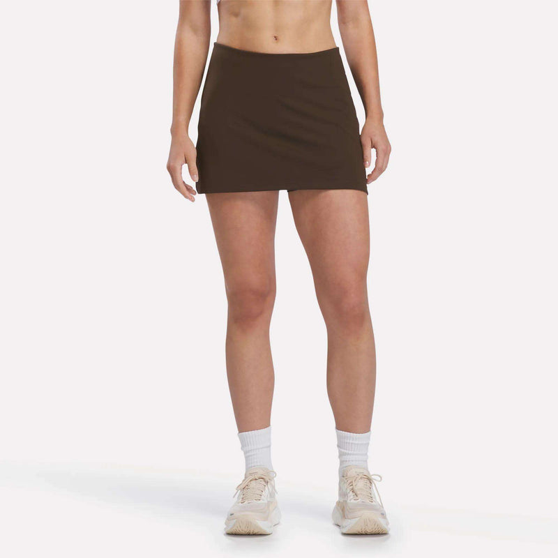 Model is shown from the waist down wearing the Reebok Women’s Tennis Skort in Espresso, paired with white socks and white sneakers, against a plain white background.