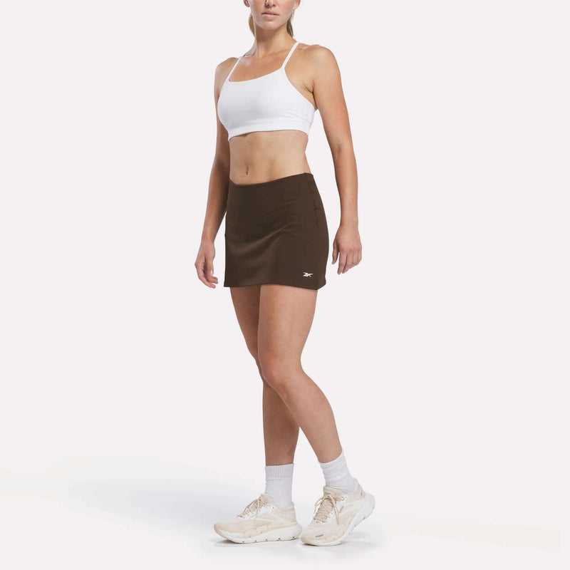 A woman stands against a plain white background, showcasing a court-inspired style in the Reebok Women’s Tennis Skort, paired with a white sports bra, white socks, and white sneakers.