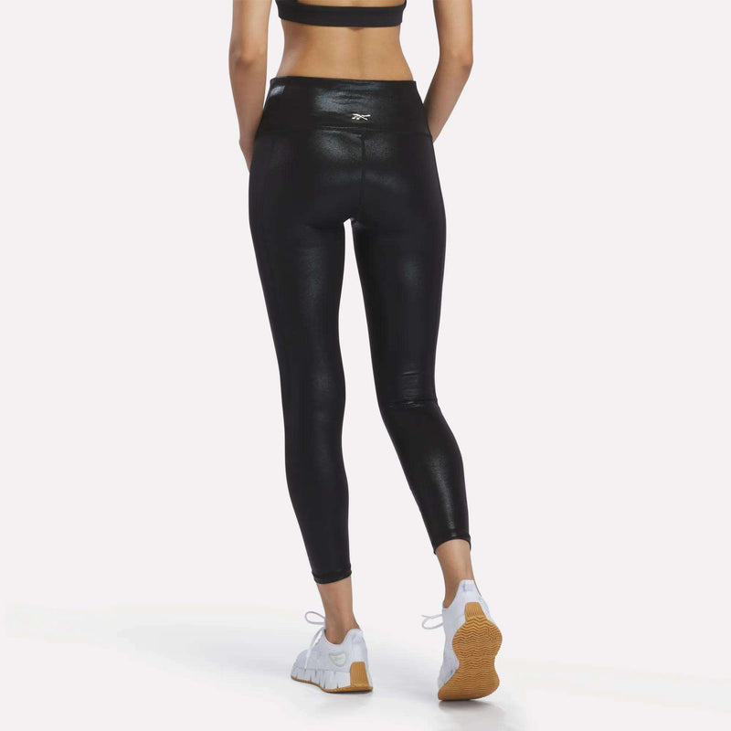 A person is shown from the back, walking forward against a plain light background, wearing black shiny Reebok Women’s Chrome 7/8 Leggings, a black sports bra, and white sneakers.