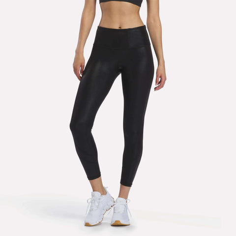 A person wearing Reebok Women’s Chrome 7/8 Leggings in black, which feature moisture-managing technology, along with a black sports bra and white athletic shoes stands against a plain white background. The person’s face is not visible in the image.