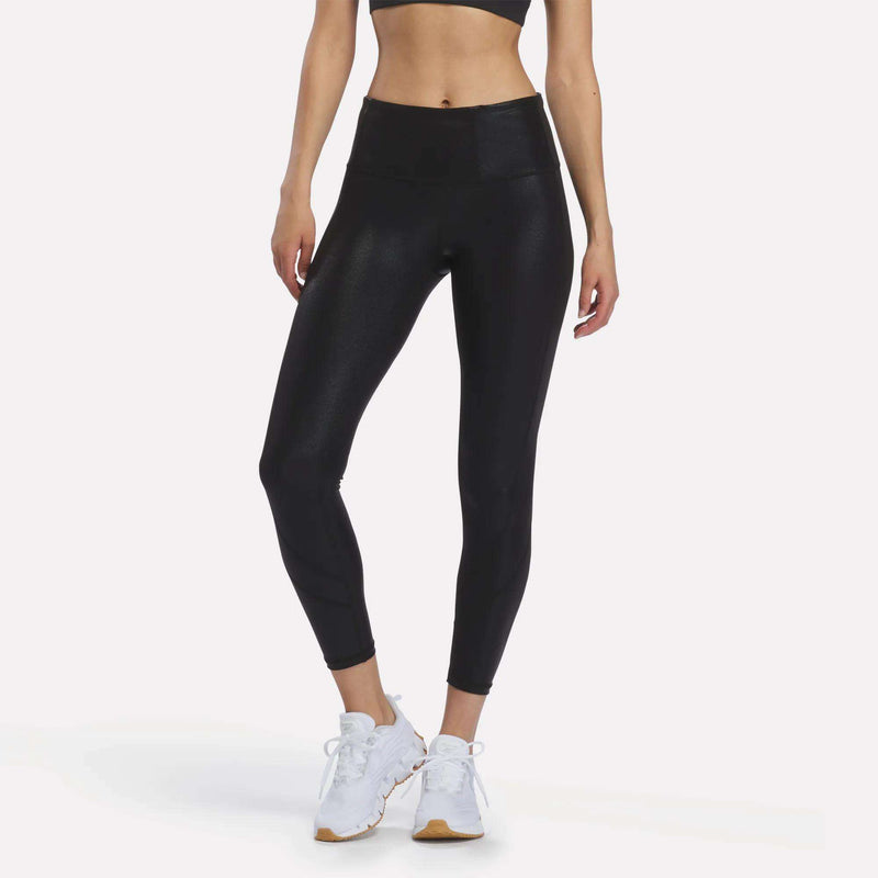 A person wearing Reebok Women’s Chrome 7/8 Leggings in black, which feature moisture-managing technology, along with a black sports bra and white athletic shoes stands against a plain white background. The person’s face is not visible in the image.