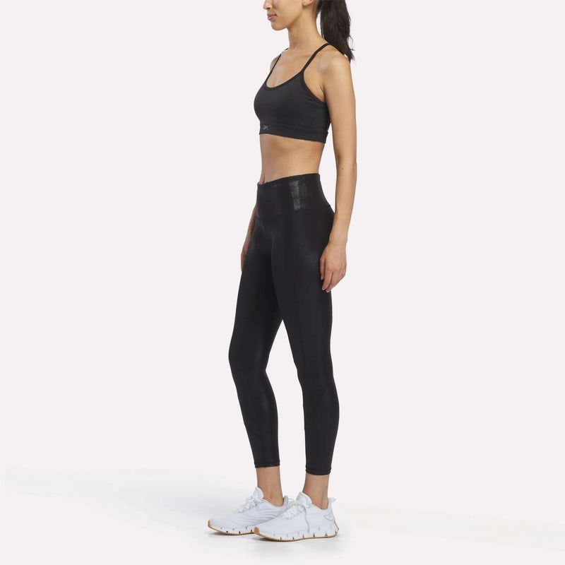 A woman in a black sports bra and Reebok Women’s Chrome 7/8 Leggings stands in profile against a white background, wearing white athletic shoes.