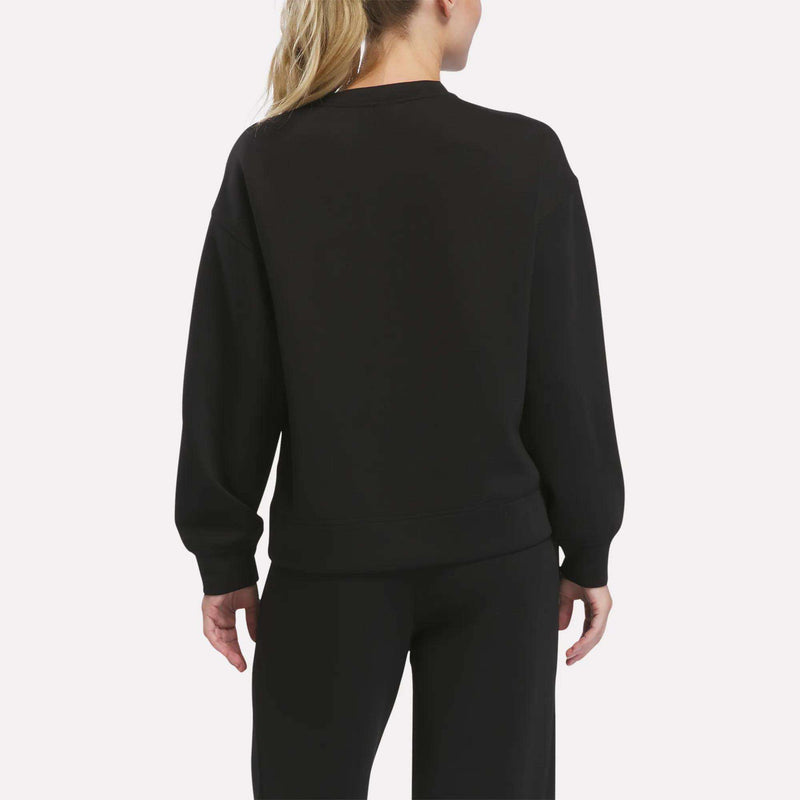 A woman with blonde hair in a ponytail is seen from behind, wearing the Reebok Women’s Crewneck Sweatshirt in black along with matching black pants, standing against a plain light background.