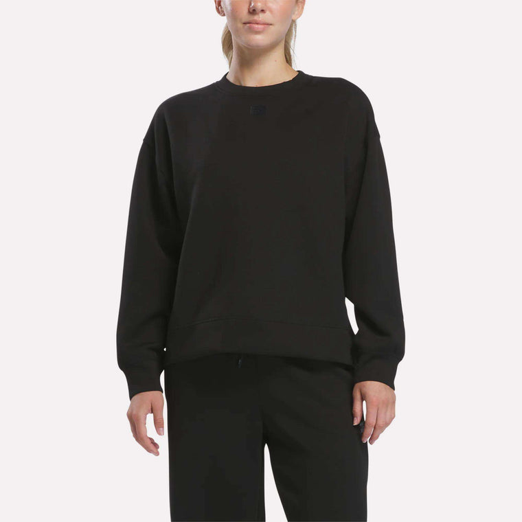 A woman wearing a loose-fitting black Reebok Women’s Crewneck Sweatshirt stands against a plain white background.