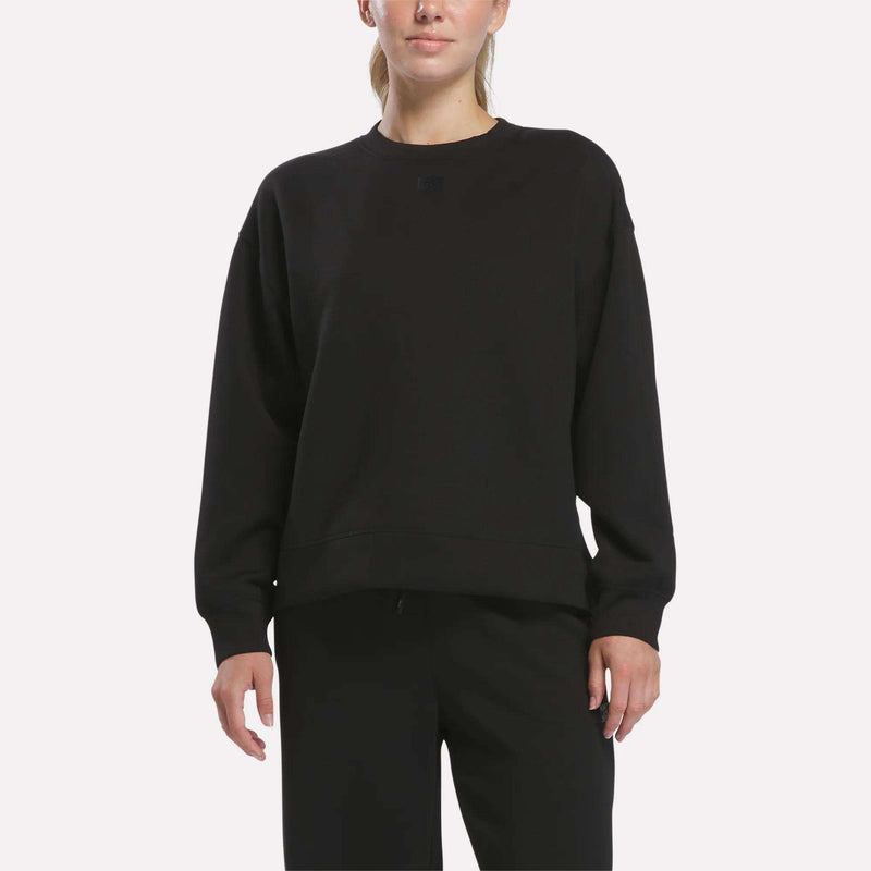 A woman wearing a loose-fitting black Reebok Women’s Crewneck Sweatshirt stands against a plain white background.
