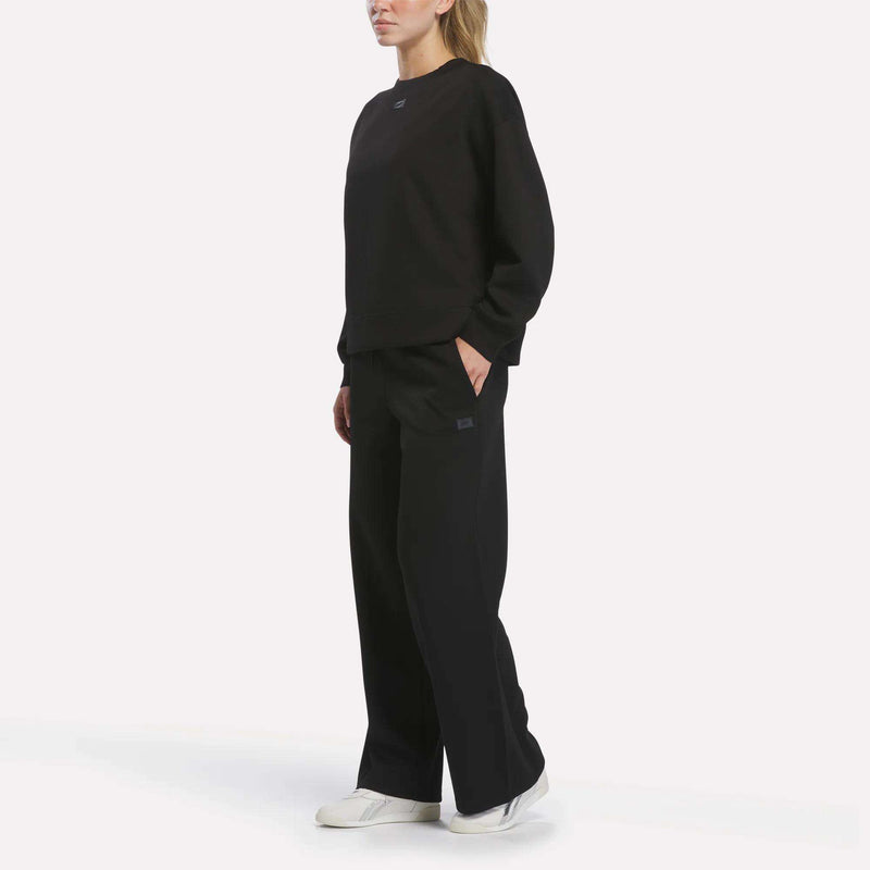 A person stands with hands in their pockets, wearing a loose-fit Reebok Women’s Crewneck Sweatshirt in black and wide-leg black pants, paired with white sneakers. The background is plain white, and their face is partially out of frame.