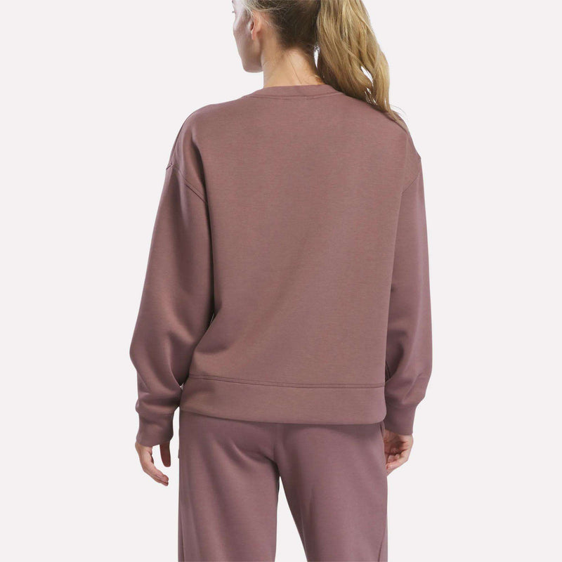 A person with long blonde hair in a ponytail is seen from behind, wearing the Reebok Women’s Crewneck Sweatshirt and matching pants in mauve, standing against a plain background.