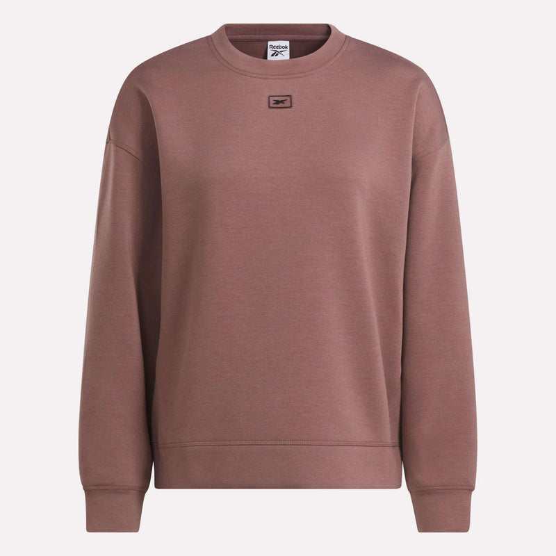 The Women’s Crewneck Sweatshirt by Reebok showcases a classic brown design for women, featuring long sleeves, ribbed cuffs, and a small rectangular logo patch on the upper chest. Made from soft doubleknit fabric, it delivers both comfort and style when worn against a plain, light-colored background.