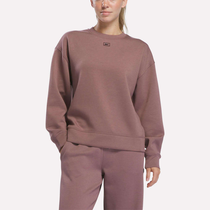 A person wearing a Reebok Women’s Crewneck Sweatshirt in Light Cocoa, along with matching pants, stands with one hand in their pocket against a plain light background.