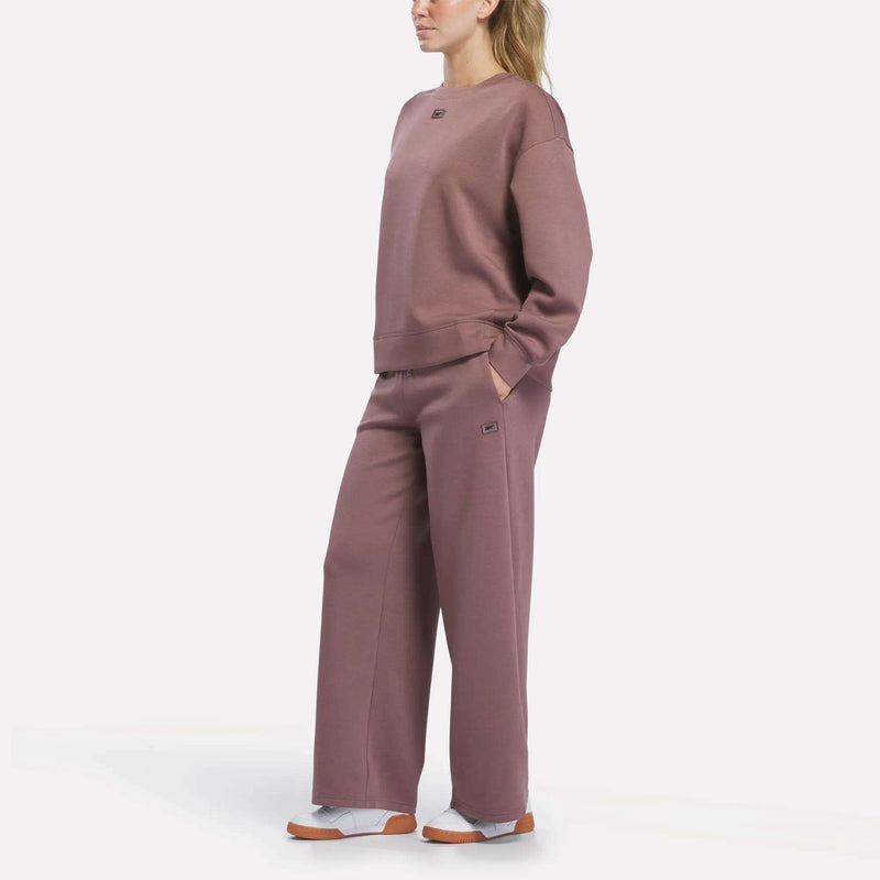 A person stands against a plain white background, hands in pockets, wearing a loose-fitting mauve Reebok Women’s Crewneck Sweatshirt paired with matching wide-leg pants and white sneakers.
