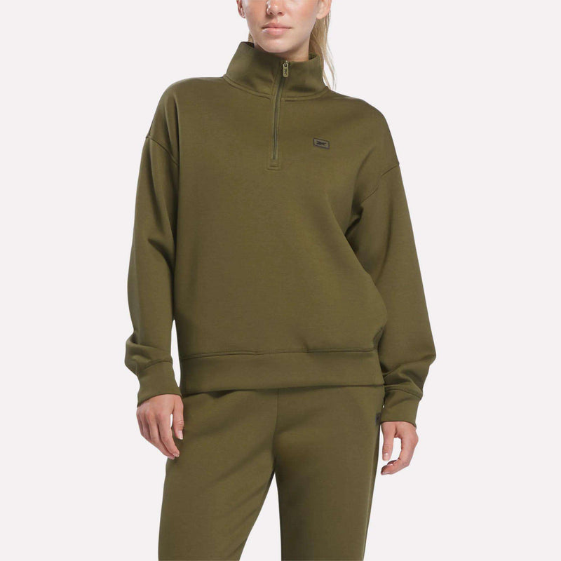 A person is wearing an olive stone Women’s Crop Half-Zip Sweatshirt by Reebok, paired with matching sweatpants, standing against a plain light background. Their hair is pulled back, and their left hand rests by their side.