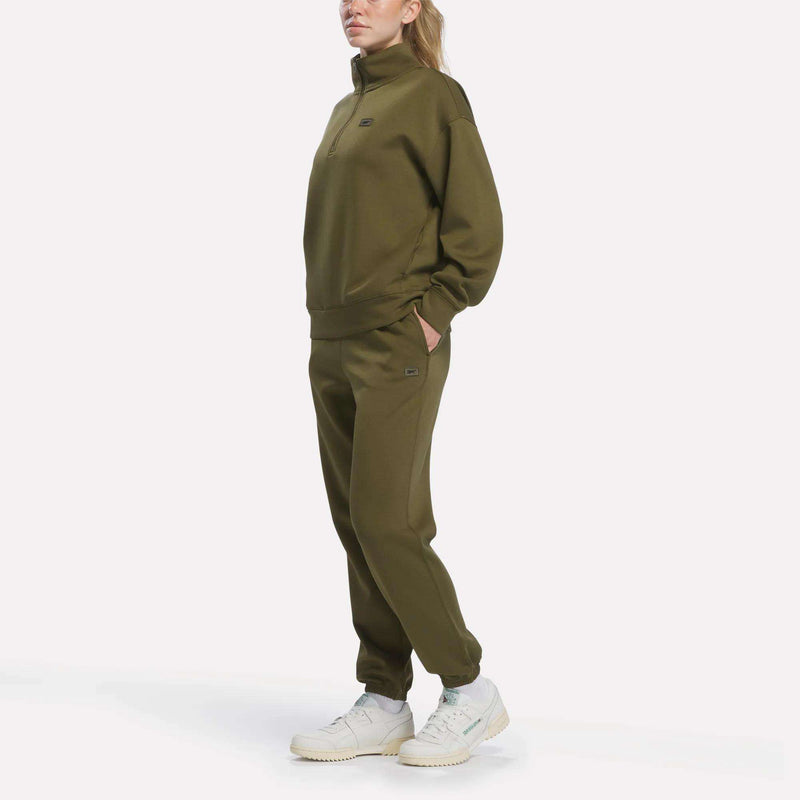 A woman stands in an olive green tracksuit with her hands in her pockets, wearing a Reebok Women’s Crop Half-Zip Sweatshirt and white sneakers against a plain white background.