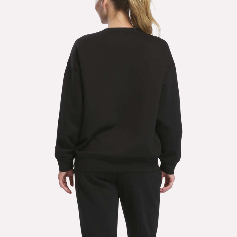 A person with blond hair in a ponytail is standing with their back to the camera against a plain white background, showcasing a relaxed silhouette while wearing the Reebok Women’s Relaxed Crewneck Sweatshirt and black pants.