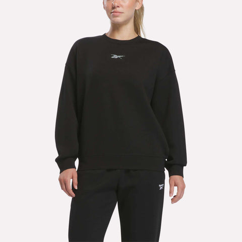 A person stands against a plain light background dressed in the Reebok Women’s Relaxed Crewneck Sweatshirt in black and matching black pants, both adorned with subtle white Reebok logos. The relaxed silhouette of the sweatshirt enhances the effortlessly stylish vibe.