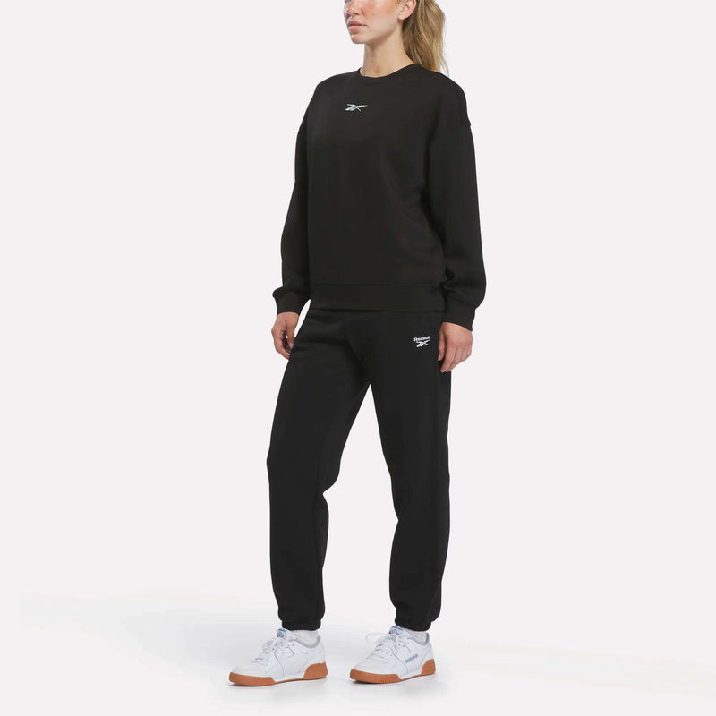 Wearing the Reebok Women’s Relaxed Crewneck Sweatshirt in black with matching sweatpants and white sneakers, a person stands against a plain white background.