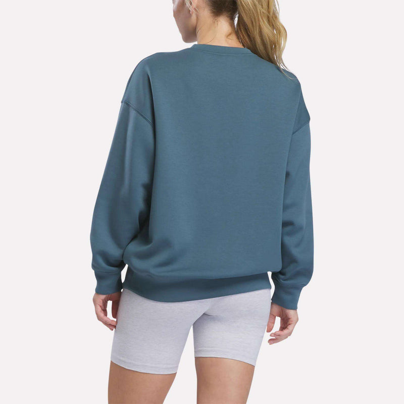 Seen from behind, a person with a ponytail wears the Reebok Women’s Relaxed Crewneck Sweatshirt paired with light gray fitted shorts, set against a plain white background to highlight the sweatshirt’s roomy silhouette.