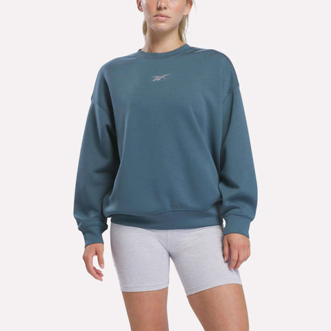 A person stands against a plain background wearing the Reebok Women’s Relaxed Crewneck Sweatshirt in Midnight Tide, which features a relaxed silhouette and a small logo on the chest, paired with light gray shorts.